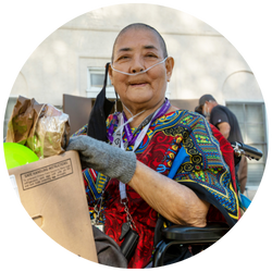 An elderly person sits in a wheelchair, oxygen tube gently curving from their nose. Dressed in a colorful outfit, they cradle a cardboard box from the Los Angeles Regional Food Bank. Festive balloons and a bag fill the background with an air of celebration.