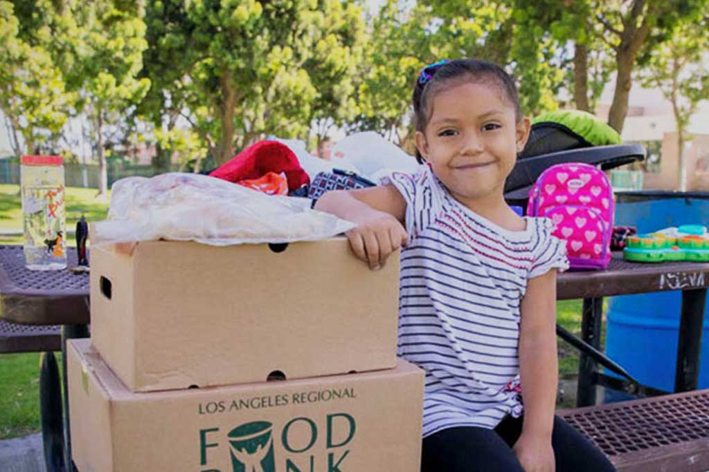 A young girl smiles while leaning on a stack of food boxes from the Los Angeles Regional Food Bank at a park. She is wearing a striped shirt with a pink hair bow, and there is a colorful backpack and trees in the background.