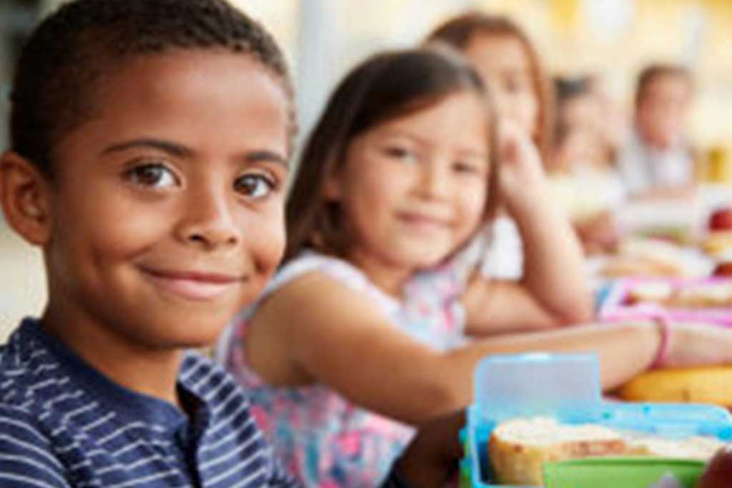 Two children sit at a table, grinning with lunch boxes from the Los Angeles Regional Food Bank before them. Other kids are blurred in the background, but the spotlight is on the boy with curly hair and his cheerful companion beside him.