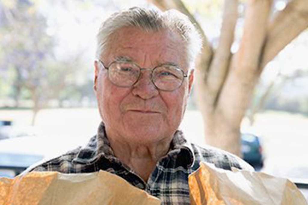 An elderly man with glasses and a plaid shirt stands outdoors clutching a brown paper bag, which he recently received from the Los Angeles Regional Food Bank. The background showcases blurred trees under bright sunlight, highlighting the warmth of community support.