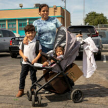 A woman stands beside a young boy and a toddler in a stroller outside the Los Angeles Regional Food Bank. They are in a parking lot with cars and a building behind them. The toddler holds a box in the stroller's basket, while various items hang from the handle.