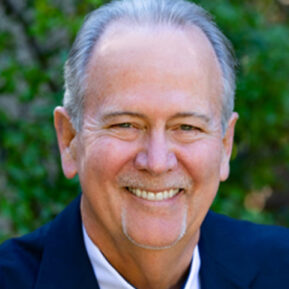 A smiling man with short gray hair and a goatee, donned in a dark suit jacket, stands outdoors against a blurred green backdrop. His commitment to the Los Angeles Regional Food Bank shines through his warm demeanor.