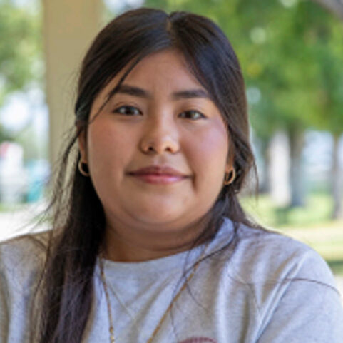 A person with long dark hair wearing a light gray shirt stands outdoors, near the Los Angeles Regional Food Bank. The background showcases green trees and soft, natural light.