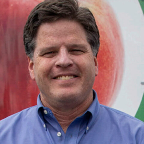 A man with dark hair wearing a blue collared shirt smiles at the camera, standing in front of the Los Angeles Regional Food Bank. A blurred image of a large fruit can be seen in the background, highlighting his commitment to nourishing communities.