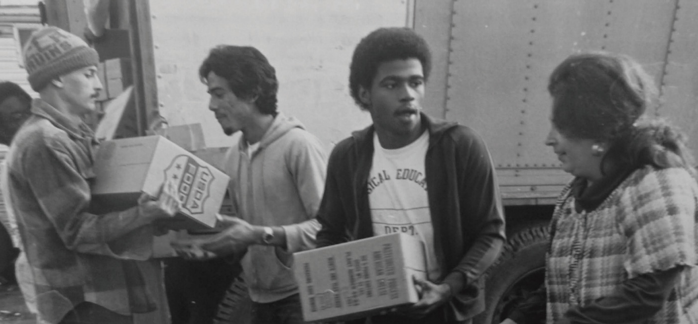 A group of people, including three men and a woman, are unloading boxes labeled "food" from a truck. They seem to be working together in a food distribution effort, possibly for the Los Angeles Regional Food Bank. The scene is captured beautifully in black and white.