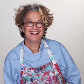 A person with curly hair and glasses is smiling, wearing a light blue shirt and a colorful, paint-splattered apron against a plain background, as they volunteer their art skills for the Los Angeles Regional Food Bank.