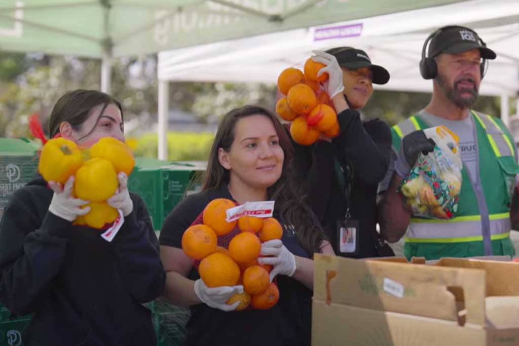 Four people under a tent hold fresh produce: two with oranges, one with bell peppers, and another with a bag of mixed fruits and vegetables. They stand behind a table with boxes, wearing gloves and casual clothes, supporting the Los Angeles Regional Food Bank initiative.