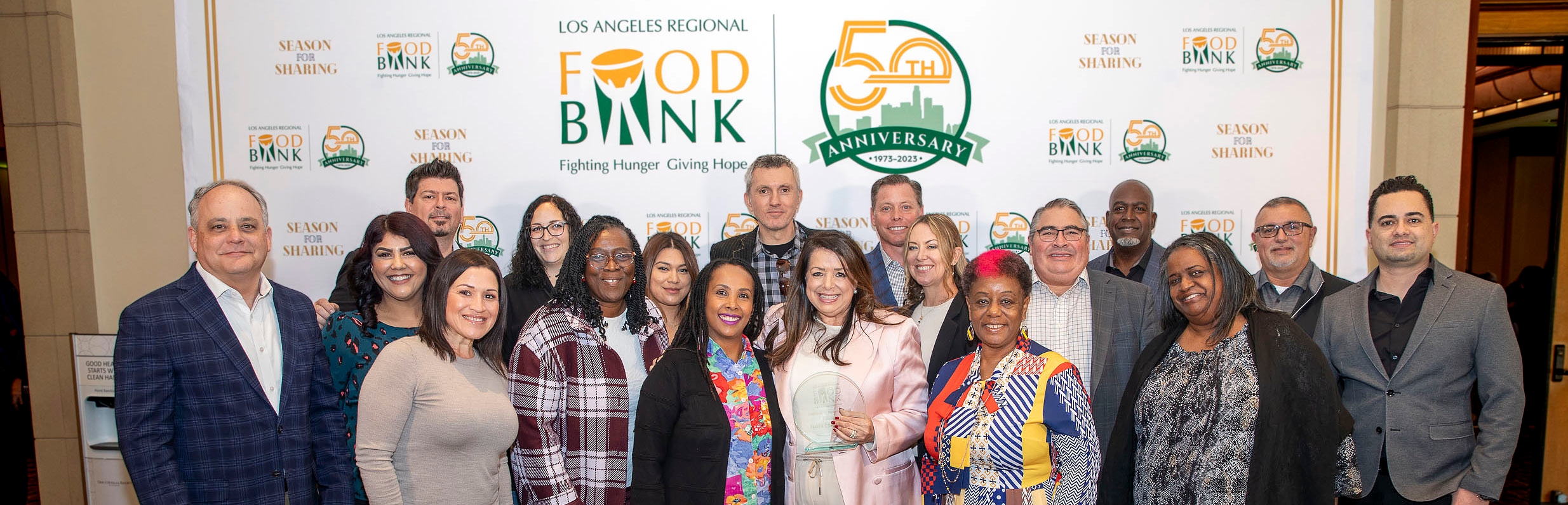 A diverse group beams with pride in front of the "Los Angeles Regional Food Bank" banner, celebrating its remarkable 50th anniversary. They proudly display an award, their smiles reflecting the profound impact of this vital organization.