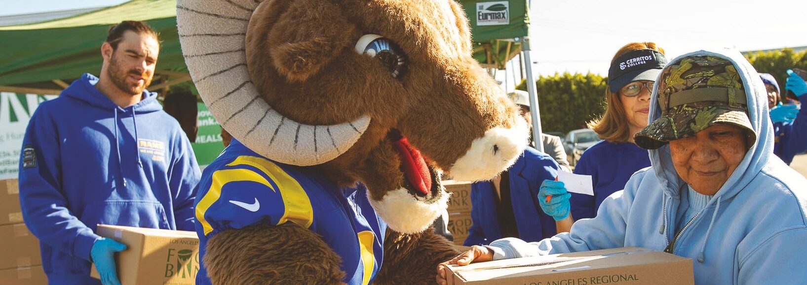 In front of a green tent, a person in a Ram mascot costume hands a Los Angeles Regional Food Bank box to an elderly person wearing a blue hoodie and camouflage cap. Other volunteers in blue shirts assist enthusiastically in the background.