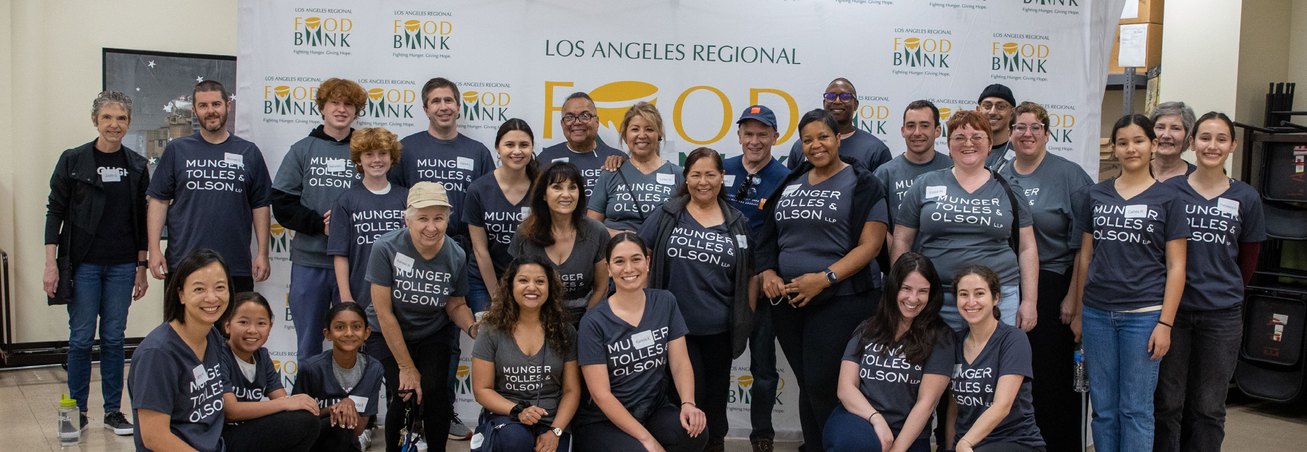 A group of diverse people, proudly donning matching shirts, poses joyfully in front of the "Los Angeles Regional Food Bank" banner. With some kneeling in the front and others standing behind them, their cheerful smiles radiate a shared sense of purpose and community spirit.