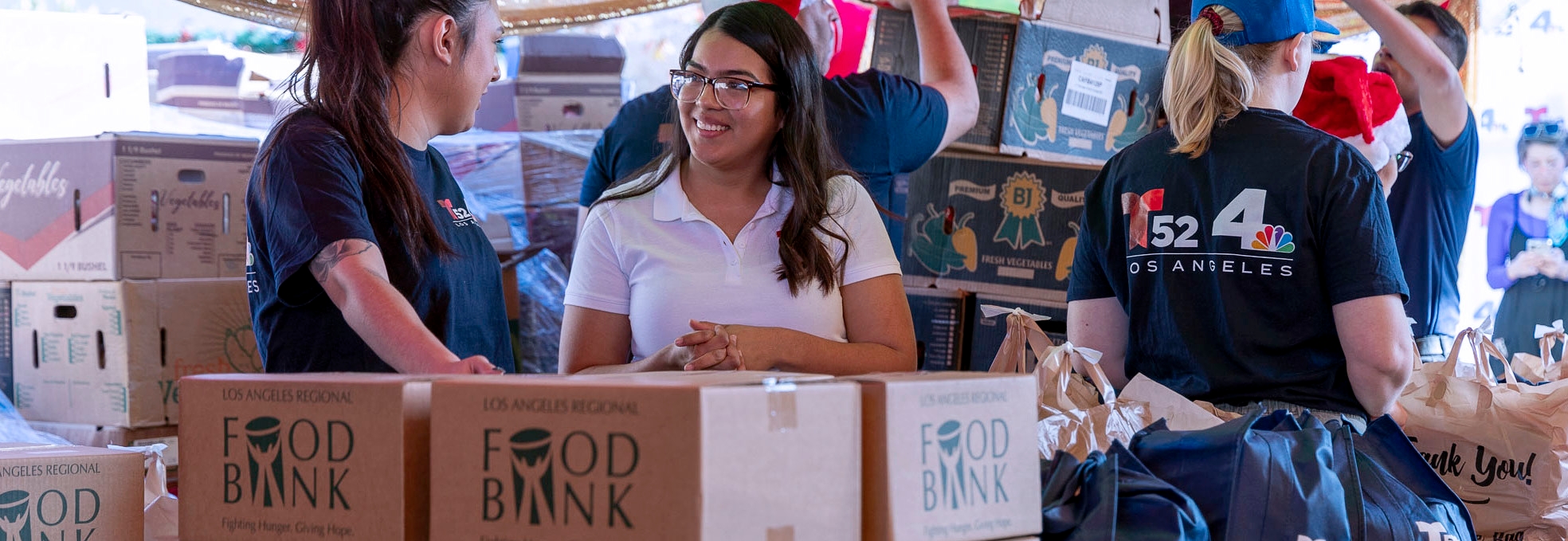 Volunteers at the Los Angeles Regional Food Bank are busy organizing boxes. Two women in the foreground chat and smile amidst stacks of cardboard boxes labeled "Food Bank." In the background, more people join in to help with this vital community effort.