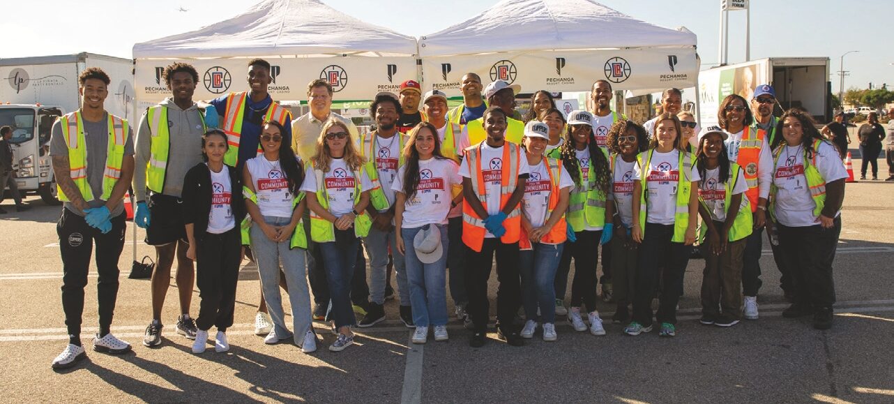 A group of diverse volunteers from the Los Angeles Regional Food Bank, wearing safety vests and gloves, stand in front of white tents at an outdoor event. Some hold bags, while others have arms around each other, smiling at the camera against a backdrop of trucks and streetlights.