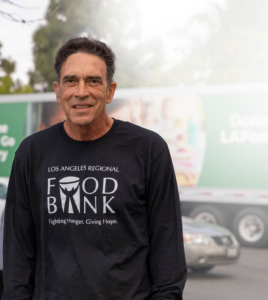 A man sporting a "Los Angeles Regional Food Bank" shirt stands outside, beaming with pride. Behind him, a truck featuring the Los Angeles Regional Food Bank advertisement completes the heartwarming scene.