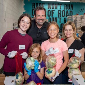 A group of five people, including two adults and three children, smile while holding bags of food at the Los Angeles Regional Food Bank. Volunteering in a facility with shelves stocked with supplies, their hands are gloved and name tags displayed. A wall sign reads "Hope Won't Run Out.