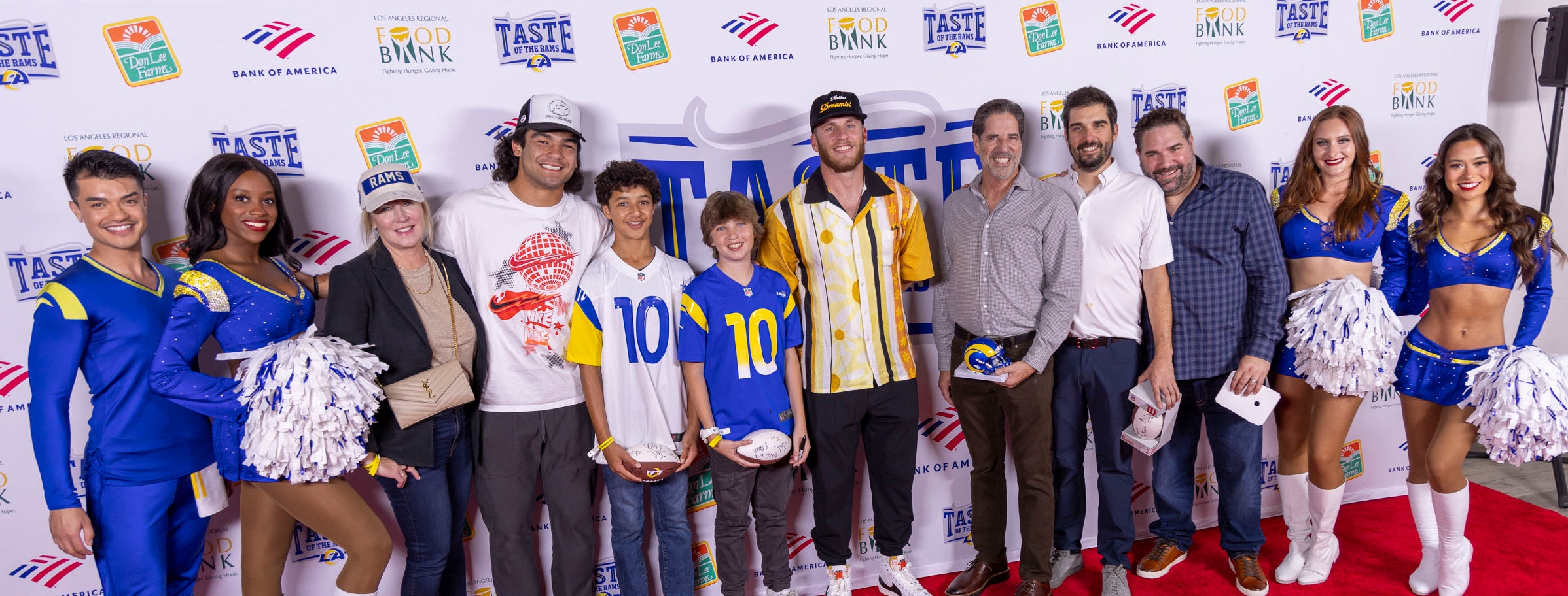 A group of people, including cheerleaders and men in casual attire, pose for a photo on a red carpet. Some hold helmets and footballs. The backdrop showcases logos like "Taste of the NFL" and "Los Angeles Regional Food Bank." Two young boys in sports jerseys add to the lively scene.