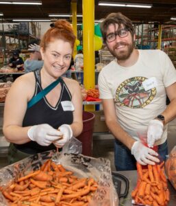 Two people happily sort and bag carrots in a bustling Los Angeles Regional Food Bank warehouse. They are wearing gloves and name tags, with other volunteers working in the background. The atmosphere is lively and cooperative as everyone contributes to a worthy cause.