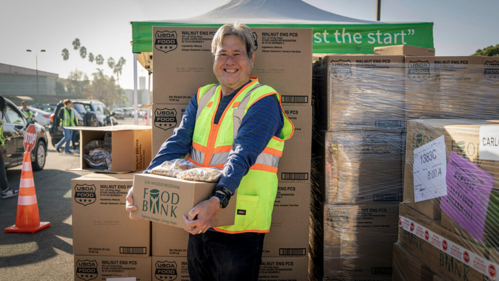 A person wearing a safety vest smiles while holding a box labeled 