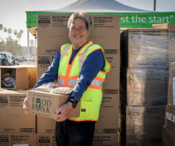 A person wearing a safety vest smiles while holding a box labeled 