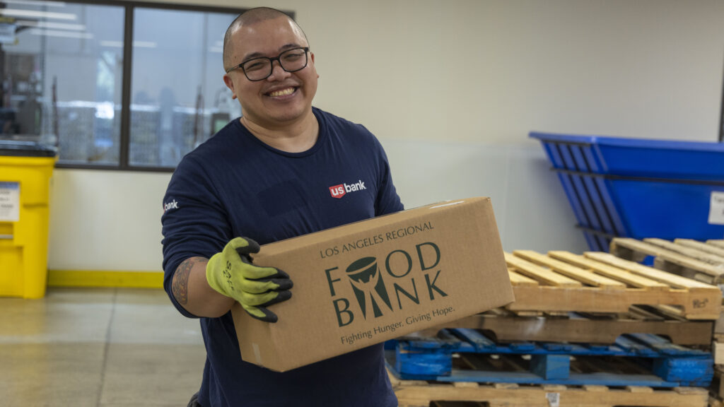 A smiling person with glasses and green gloves, representing the spirit of US Bank, holds a cardboard box labeled 