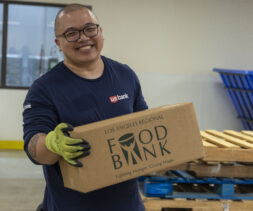 A smiling person with glasses and green gloves, representing the spirit of US Bank, holds a cardboard box labeled 