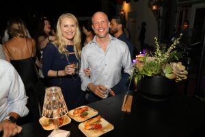 A smiling couple stands together at a social event hosted by the Los Angeles Regional Food Bank, holding drinks. They are in a lively setting with other guests in the background. A small table nearby displays plates of food and a decorative plant.