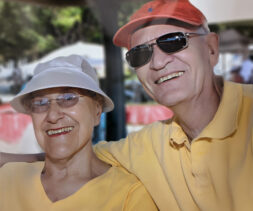 An elderly couple in matching yellow shirts and hats, one white and one red, smiles as they relax outdoors. Both wear sunglasses, enjoying a sunny day under the shade. The setting, with trees and a blurred background, hints at a park or garden—a serene moment reflecting the joy of estate giving.