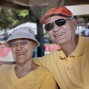 An elderly couple in matching yellow shirts and hats, one white and one red, smiles as they relax outdoors. Both wear sunglasses, enjoying a sunny day under the shade. The setting, with trees and a blurred background, hints at a park or garden—a serene moment reflecting the joy of estate giving.