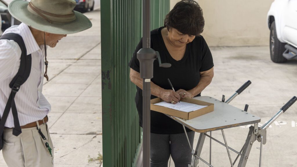 A woman writes on a clipboard resting on a makeshift table outdoors, possibly organizing for the local food bank. A man with a hat and backpack stands nearby, observing the clipboard. They are positioned on a paved area beside a green fence and a parked white vehicle.