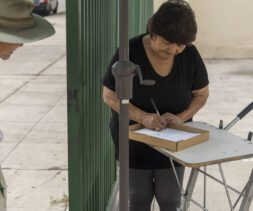 A woman writes on a clipboard resting on a makeshift table outdoors, possibly organizing for the local food bank. A man with a hat and backpack stands nearby, observing the clipboard. They are positioned on a paved area beside a green fence and a parked white vehicle.