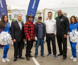 A group of six people, four men in casual and business attire and two women in blue cheerleader uniforms, pose together outdoors at a food bank event. Some hold white pom-poms. Banners and a truck are visible in the background.