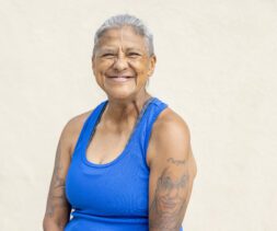 A smiling older woman with short gray hair and tattoos on her arms stands before a plain, light-colored background. Clad in a bright blue sleeveless shirt, she exudes warmth and confidence, embodying a door of hope for those who see beyond the surface.