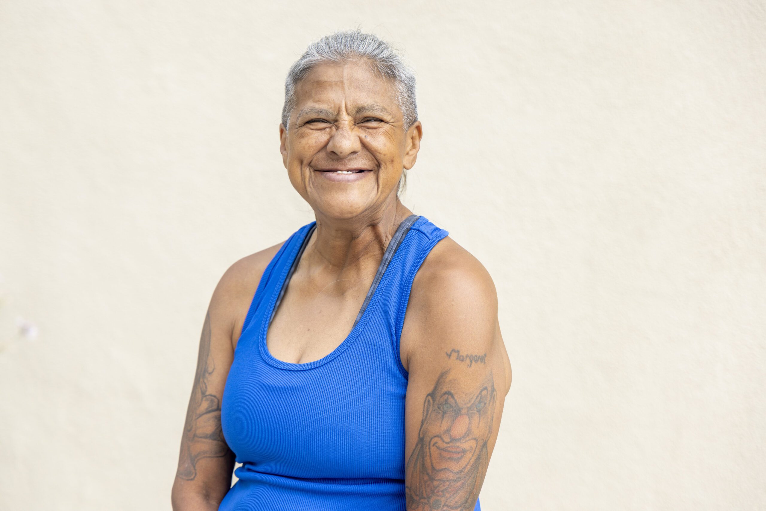 A smiling older woman with short gray hair and tattoos on her arms stands before a plain, light-colored background. Clad in a bright blue sleeveless shirt, she exudes warmth and confidence, embodying a door of hope for those who see beyond the surface.