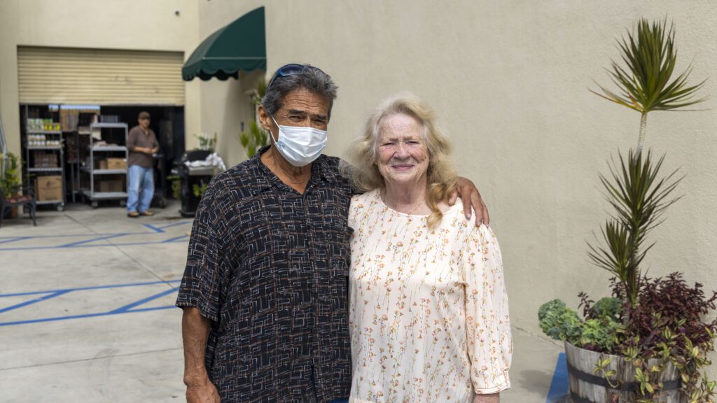 A man wearing a patterned shirt and mask stands with his arm around a smiling woman in a floral dress. They pose outdoors beside a plant, near carts and shelves at the bustling food pantry, with a wall and another person in the background.