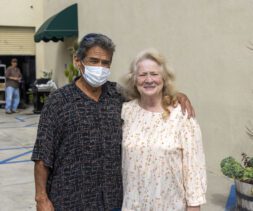 A man wearing a patterned shirt and mask stands with his arm around a smiling woman in a floral dress. They pose outdoors beside a plant, near carts and shelves at the bustling food pantry, with a wall and another person in the background.