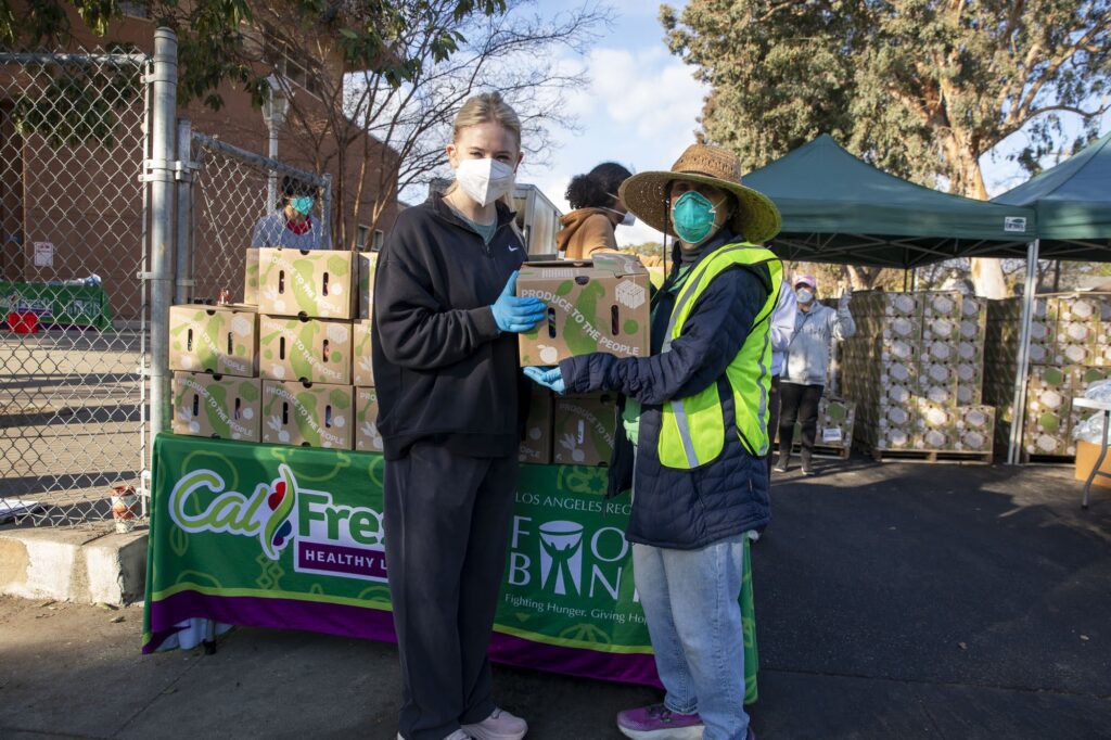 Two volunteers wearing masks distribute boxes of food outdoors at a food bank event. One person in a black outfit and another in a bright vest and hat stand in front of a table with stacked boxes and a 