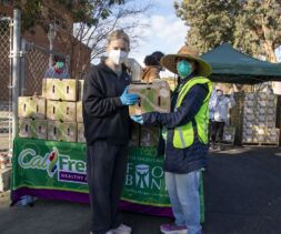 Two volunteers wearing masks distribute boxes of food outdoors at a food bank event. One person in a black outfit and another in a bright vest and hat stand in front of a table with stacked boxes and a 