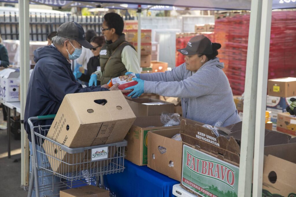 At a food distribution event, volunteers, donning masks, distribute items from boxes under a canopy. A recipient with a shopping cart collects goods, reminiscent of food pantries’ efforts. Numerous cardboard boxes are neatly arranged on tables.