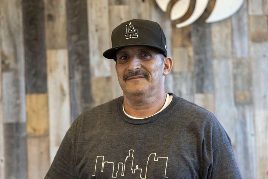 A man wearing a black cap and a dark t-shirt with a city skyline design stands in front of a wooden wall, reminiscent of the ones found at local food pantries. He has a mustache and is smiling softly at the camera.