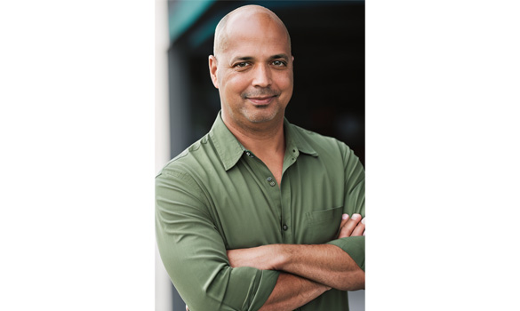 A person wearing a green button-up shirt stands with arms crossed, smiling warmly at the camera. The background is blurred, hints of blue and white suggesting an outdoor setting perhaps near a local food bank.