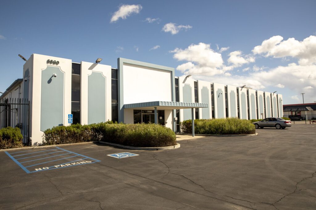 Single-story commercial building with a blue and white facade, large windows, and flat roof houses a food bank. A parking lot with reserved accessible spaces is out front under a bright sky with a few clouds.