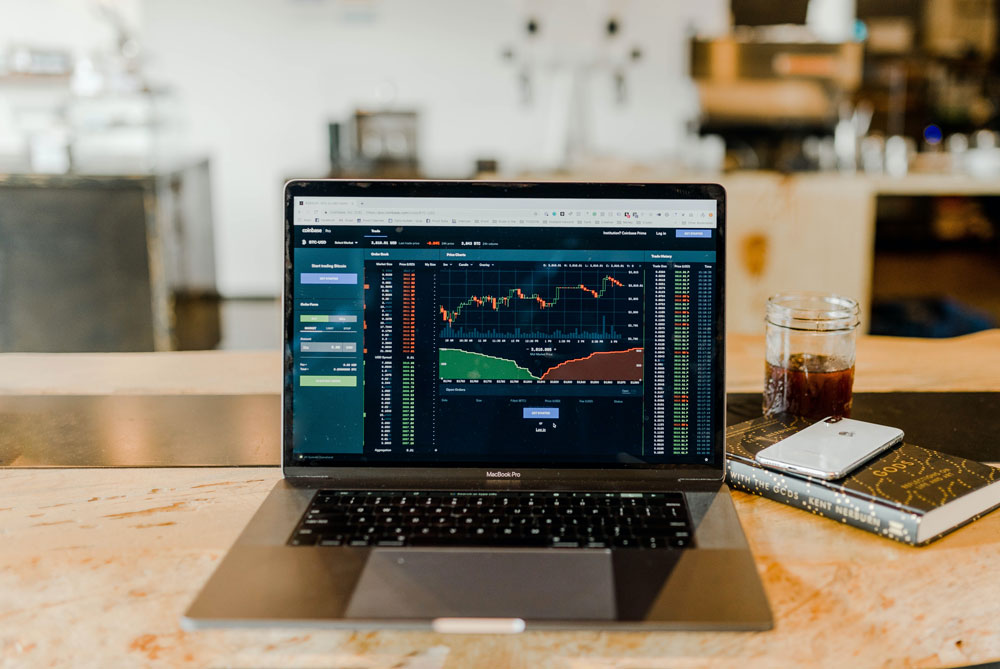 A laptop on a wooden table displays a financial trading platform with charts and graphs. Beside it, supporting the Los Angeles Regional Food Bank initiative, lies a branded notebook, alongside a smartphone and a glass of iced coffee. The background is a blurred workspace.